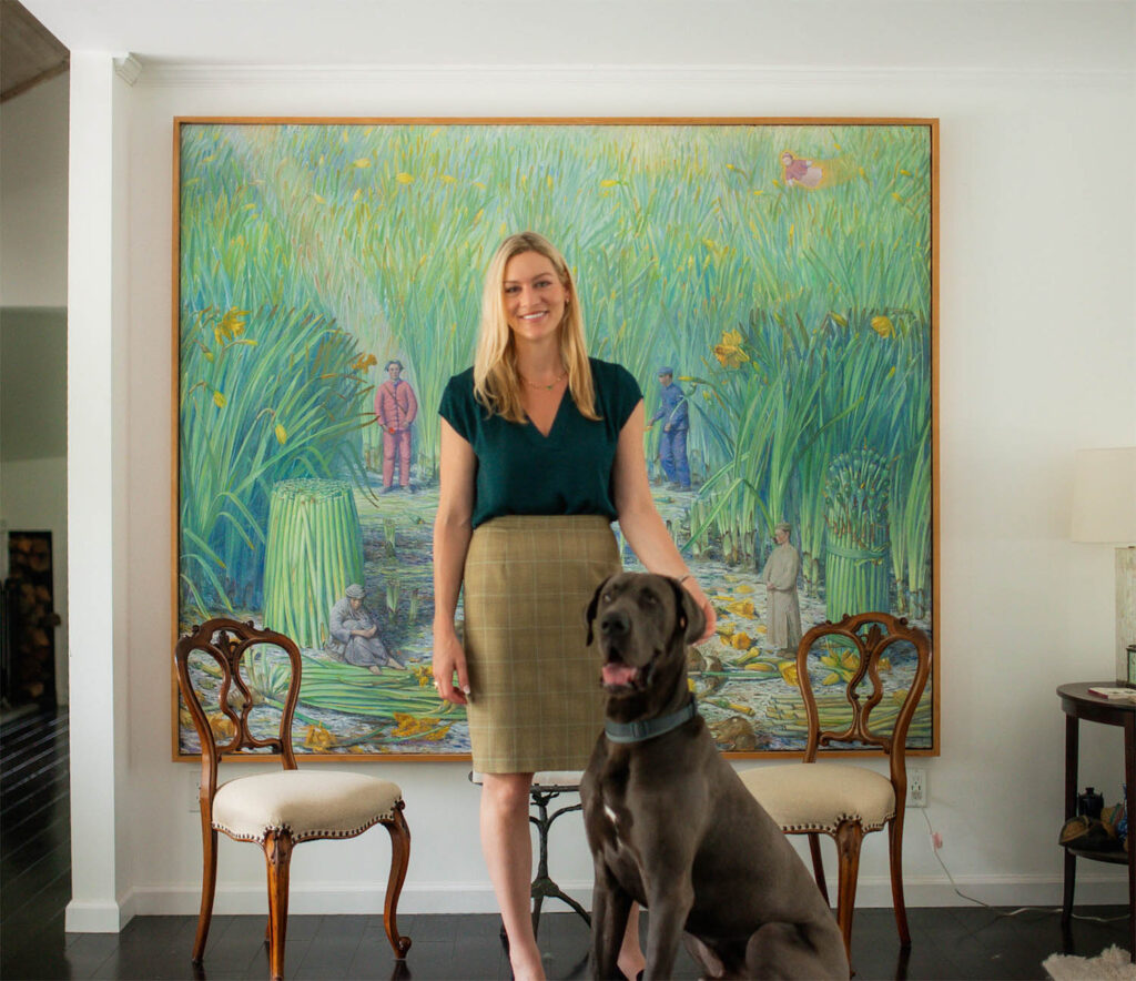A gray hair beautiful women standing with dog, and a wall painting in the background with chairs.