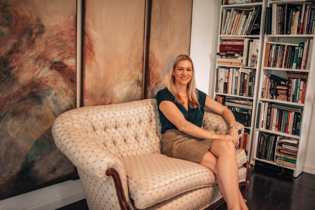 A beautiful smilling women sitting on couch, with a bookshelf on the left corner and creative painting on the background.